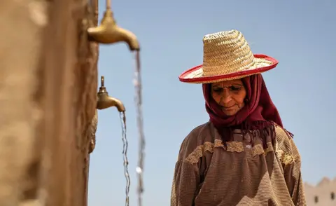 AFP A person looks down while wearing a sunhat with taps pouring water into containers (out of frame) with a blue sky in the background, in Morocco in June 2024