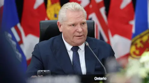 Getty Images Doug Ford speaks at a meeting in Canada. He is sitting by a desk in front of a row of national flags, including those belonging to the UK and Canada.  