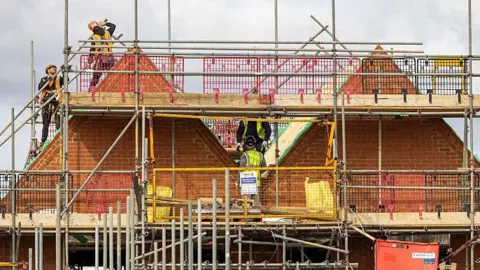 Getty Images A partially constructed brick building surrounded by extensive metal scaffolding. Several construction workers wearing safety gear are working on the upper level near triangular roof structures. The site includes wooden planks, metal poles, and safety barriers in pink, grey and yellow colours. A cloudy sky forms the background.
