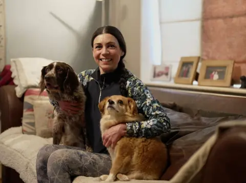 Helene Svinos sits on a sofa in between two dogs, with her arms around them. She is smiling and wearing a dark gilet over a patterned top, and leggings, with her dark hair tied back. There are cushions on the sofa and photo frames in the background.